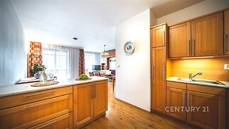 Kuchyň s dřevěnými skříňkami, parketovou podlahou a moderním designem plynule přechází do světlého obývacího pokoje s velkými okny.
