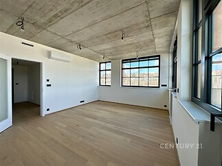 Světlý obývací pokoj s parketovou podlahou, velká okna a moderní betonový strop v rekonstruovaném bytě.