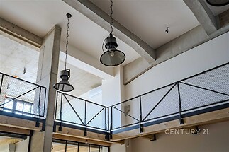 Moderní loftový prostor s vysokými stropy, betonovými prvky a kovovým zábradlím. Průmyslový styl a velká okna poskytují dostatek světla.