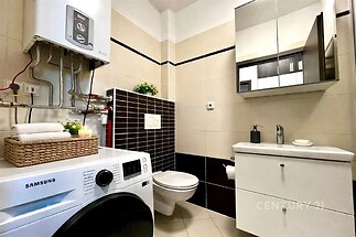Malá moderní koupelna s dlažbou, pračkou a vestavěným úložným prostorem. Světlé obklady a tmavé dekorační prvky dodávají styl.