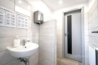 Veřejná toaleta s moderními obklady, umyvadlem a samostatným uzavřeným prostorem. Světlé dlaždičky a kovové doplňky přispívají k čistému vzhledu.