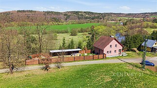 Rodinný dům s růžovou fasádou, dřevěným plotem a šikmou střechou. Nachází se na okraji lesa s výhledem na zelené louky.