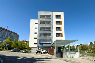 Moderní bytový dům s hlasitými liniemi, balkony a šedým obkladem, obklopený zelení, v klidné oblasti.