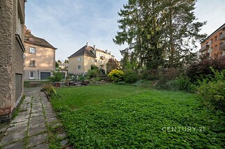 Zahrada s travnatou plochou, obklopená stromy a keři, přiléhající k rekonstruovanému domu, v klidné vilové čtvrti.