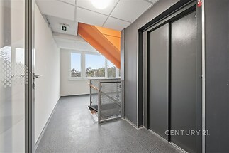 Chodba s moderním výtahem, kovovým zábradlím a velkými okny, která zajišťují dostatek denního světla, na podlaze je praktická dlažba.