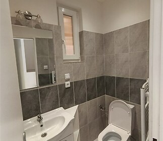 Koupelna s moderní dlažbou, umyvadlovou skříňkou a světle šedými obklady s malým oknem poskytujícím přirozené světlo.