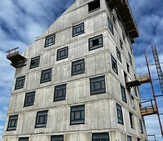 Šestipatrová betonová budova s velkými okny a moderní konstrukcí, která je součástí nové výstavby, nabízí potenciál pro rezidenční nebo komerční využití.