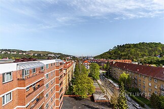 Výhled z bytu na klidnou ulici s cihlovými domy, okolní zelení a kopcovitou krajinou, ideální pro milovníky přírody a městského života.