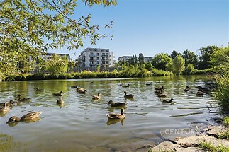 Světlé okolí bytového komplexu s výhledem na jezero a lavičky, obklopené zelení a moderní architekturou, idealní pro relaxaci v přírodě.
