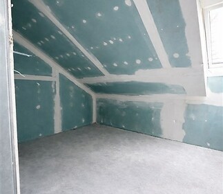 Podkrovní místnost s nedokončenými stěnami, velkým oknem a betonovou podlahou, ideální pro rekonstrukci nebo kreativní úpravy.