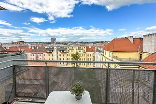 Balkon s kovovým zábradlím a výhledem na město, světlé cihlové budovy s červenými střechami, modrá obloha s mraky