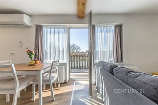 Světlý obývací pokoj s parketovou podlahou a velkými okny, které vedou na balkon. Moderní nábytek a výhled na okolní krajinu.