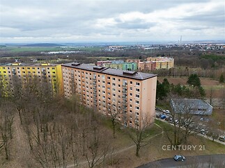 Pohled na panelový bytový dům obklopený zelení, s parkovištěm v pozadí a barevnými sousedními budovami, situovaný v klidné lokalitě.