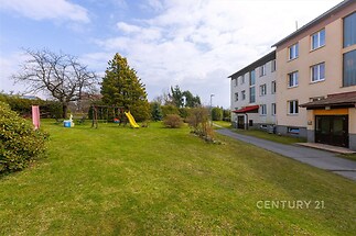 Zahrada bytového domu s dětským hřištěm, udržovaný trávník, klidné prostředí, parkovací plocha v blízkosti, vhodné pro rodiny s dětmi.