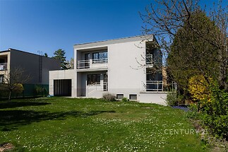 Moderní rodinný dům s bílou fasádou a velkými okny, nacházející se uprostřed prostorné zahrady s udržovaným trávníkem.