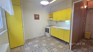 Kuchyň s retro dlažbou, žlutými skříňkami a plynovým sporákem. Prostor je světlý, se závěsem nad oknem a dřevěným obložením na stěně.