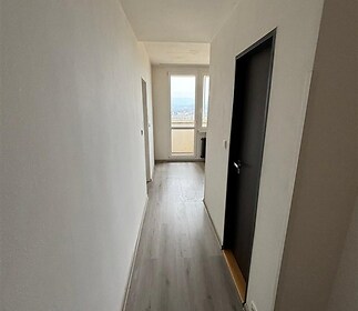 Úzká chodba s moderní dřevěnou podlahou, zakončená balkonovými dveřmi. Světlé zdi dodávají prostoru vzdušnost a umožňují přirozené světlo.
