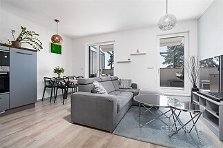 Světlý obývací pokoj s moderní sedačkou, parketová podlaha, velká okna vedoucí na balkon, moderní osvětlení, jídelní stůl pro čtyři.