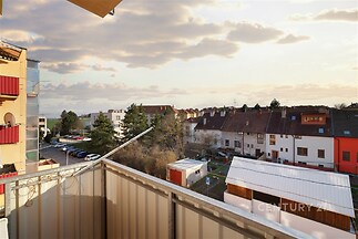 Balkón s výhledem na okolní zástavbu, moderní kovové zábradlí, příjemné odpolední světlo, klidná rezidenční čtvrť, ideální pro relaxaci.