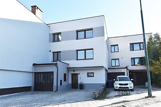 Fasáda moderního rodinného domu s bílou omítkou a šedými akcenty, garáž a dlážděná příjezdová cesta, před domem zaparkované auto.