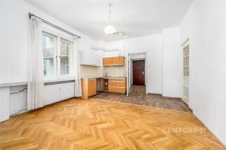 Světlý obývací pokoj s parketovou podlahou a velkými okny, propojený s kuchyní, která má moderní spotřebiče a dlažbu v elegantním designu.