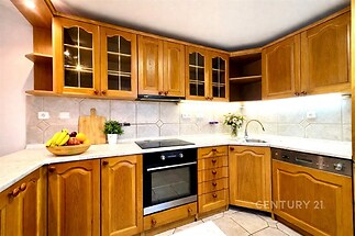 Kuchyň s dřevěnými skříňkami, moderní spotřebiče, světlo z velkých oken, dlažba na podlaze, světlé dekorace, kvalitní pracovní deska.