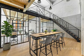 Moderní kancelářský prostor s dřevěnou podlahou, skleněnými příčkami, a ocelovým schodištěm. Velká okna poskytují dostatek přirozeného světla.