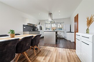 Moderní kuchyň s parketovou podlahou, velkými okny a vestavěnými spotřebiči. Světlý otevřený prostor s dřevěným jídelním stolem a světlým interiérem.