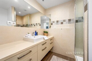 Moderní koupelna s dlažbou, velkým zrcadlem a sprchovým koutem. Světlý interiér a elegantní úložné skříňky dodávají prostoru stylový vzhled.