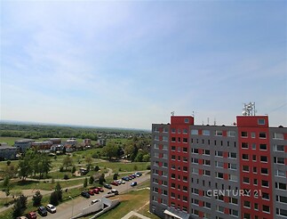 Výhled z balkonu na moderní bytový dům s červenými detaily, okolní zeleň a parkoviště, světlý prostor s výhledem na krajinu