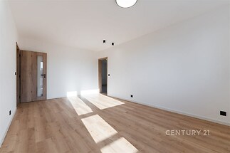 Světlý obývací pokoj s parketovou podlahou, velká okna poskytují dostatek přirozeného světla, moderní styl a neutrální barvy stěn.