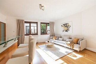 Světlý obývací pokoj s parketovou podlahou, velkými okny a moderním nábytkem, nabízí pohodlné posezení a elegantní designový styl.