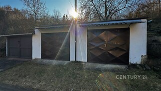 Dvojgaráž s kovovými vraty, rovná střecha a betonová konstrukce, nachází se na klidném místě obklopeném zelení, ideální pro parkování nebo skladování.
