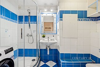 Koupelna s modro-bílými obklady, sprchovým koutem a umyvadlem. Prostředí je čisté, moderní a obsahuje zabudovanou pračku.
