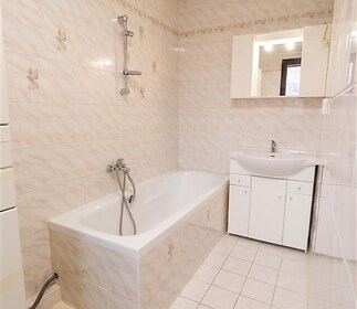 Koupelna s dlažbou a obklady, vana s ruční sprchou, světlý interiér, moderní umyvadlová skříňka a zrcadlo s úložným prostorem.