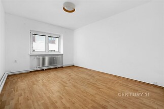 Světlý pokoj s parketovou podlahou, bílými stěnami a velkým oknem, který poskytuje dostatek přirozeného světla, ideální pro moderní bydlení.