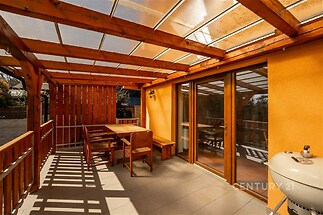 Zastřešená terasa s dřevěným nábytkem a velkými okny nabízí moderní design a kvalitní dlažbu, ideální pro venkovní posezení.