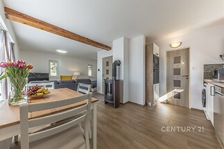 Světlá jídelna s dřevěným stolem, parketová podlaha, moderní spotřebiče, krbová kamna a velká okna dodávají prostoru moderní vzhled.