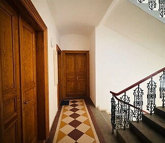 Chodba v historickém stylu s dlažbou, dřevěnými dveřmi a kovovým zábradlím na schodech, evokující atmosféru klasického činžovního domu.