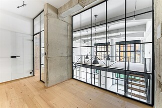 Průmyslový interiér s moderními prvky, velká okna a skleněné příčky, parketová podlaha, světlý prostor s betonovými prvky.