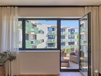 Světlý obývací pokoj s velkými okny, parketovou podlahou a moderním balkonem nabízí výhled na sousední budovy.