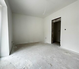 Prázdná místnost s betonovou podlahou a bílými stěnami, připravená k dokončení. Přirozené světlo z velkého okna, základní prostor pro úpravy.