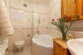 Koupelna s rohovou vanou, dlažbou a dřevěným nábytkem, světlé obklady a moderní sprchový kout vytvářejí příjemný a praktický prostor.