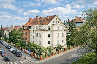 Klasický bytový dům s červenou střechou, nacházející se na rohu klidné městské ulice, obklopený zelení a historickými budovami.