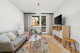 Světlý obývací pokoj s parketovou podlahou, velká okna nabízejí přírodní světlo, moderní nábytek v minimalistickém stylu.