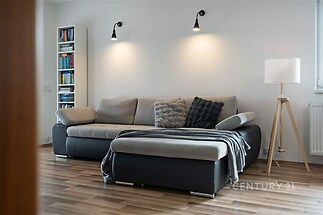 Moderní obývací pokoj, parketová podlaha, pohodlná sedací souprava, světlé stěny, knihovna, designové osvětlení.