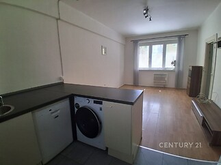 Světlý obývací pokoj s parketovou podlahou a velkými okny, moderní kuchyňský kout s vestavěnými spotřebiči, celkově moderní uspořádání.