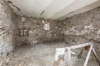 Prázdný prostor se surovými kamennými zdmi, hrubou betonovou podlahou a malým oknem, ideální pro rekonstrukci nebo dílenské využití.