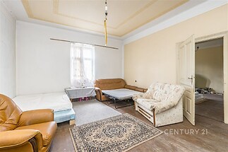 Světlý obývací pokoj s dřevěnou podlahou, velkými okny a vysokými stropy, v původním stavu s možností rekonstrukce.