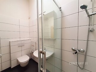 Malá koupelna s moderní dlažbou, sprchovým koutem a bílými kachličkami, vybavená závěsnou toaletou a umyvadlem s úložnou skříňkou.